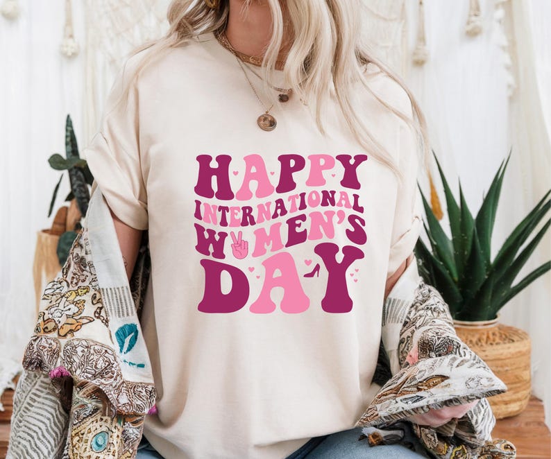 Op de afbeelding: Een wit T-shirt met de tekst "Happy International Women's Day" in roze en paarse letters. De letters zijn gestileerd met harten en een vredesteken.
