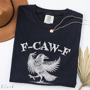 May include: Black t-shirt with a graphic of a raven and the text "F-CAW-F" in white. A gold necklace and a brown hat are also in the image. The shirt is a Comfort Colors brand.