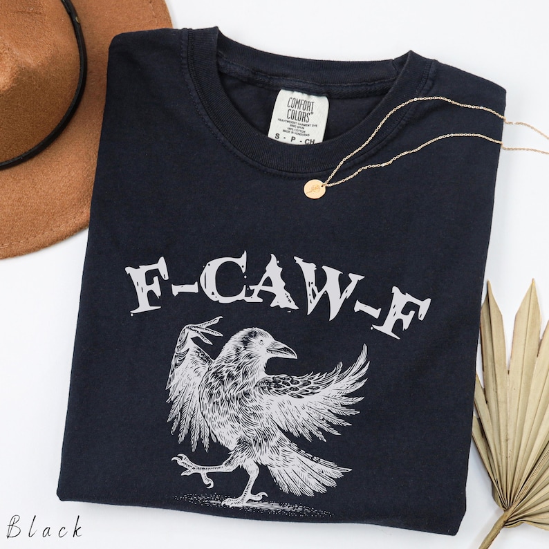 May include: Black t-shirt with a graphic of a raven and the text "F-CAW-F" in white. A gold necklace and a brown hat are also in the image. The shirt is a Comfort Colors brand.