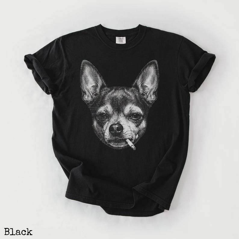 May include: Black t-shirt with a monochrome illustration of a Chihuahua smoking a cigarette. The design is a detailed drawing of the dog's face. The t-shirt has a crew neck and short sleeves. The word "Black" is printed at the bottom.