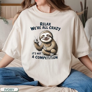 May include: A white t-shirt with a sloth graphic and the text "Relax We're All Crazy It's Not A Competition".