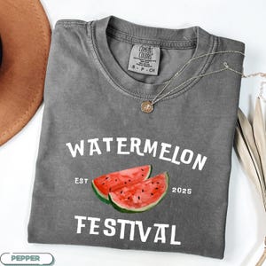 Pu&ograve; includere: Una maglietta grigia con la scritta bianca "WATERMELON FESTIVAL" e un'illustrazione ad acquerello di due fette di anguria. La maglietta ha anche il testo "EST 2025". Una collana d'argento con un ciondolo &egrave; drappeggiata sulla maglietta.