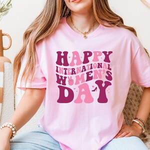 Op de afbeelding: Roze T-shirt met de tekst "Happy International Women's Day" in een retro lettertype met roze en bordeauxrode kleuren.