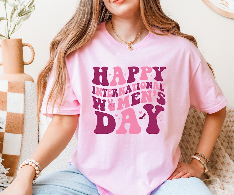 Op de afbeelding: Roze T-shirt met de tekst "Happy International Women's Day" in een retro lettertype met roze en bordeauxrode kleuren.