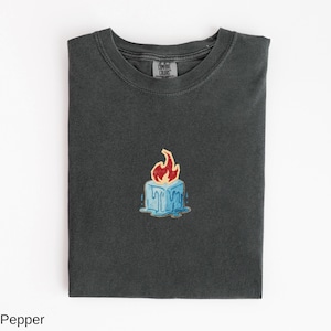 May include: Dark grey t-shirt with a small, colorful graphic of a blue ice cube with a red flame on top. The shirt is folded neatly, showing the Comfort Colors brand tag.