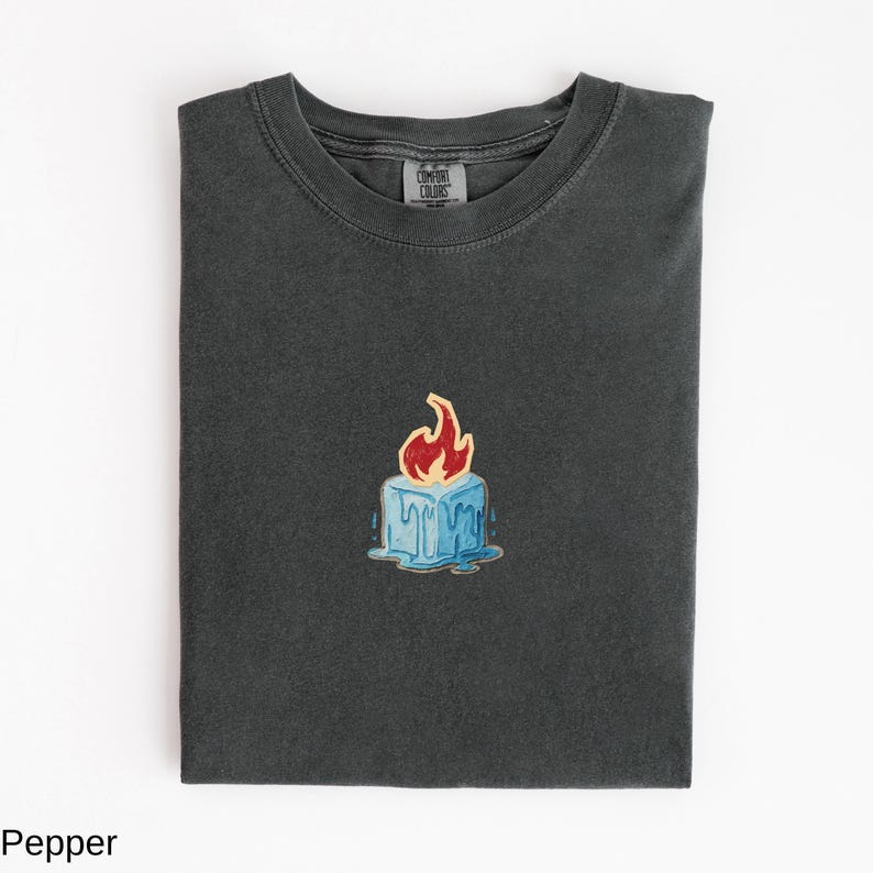 May include: Dark grey t-shirt with a small, colorful graphic of a blue ice cube with a red flame on top. The shirt is folded neatly, showing the Comfort Colors brand tag.