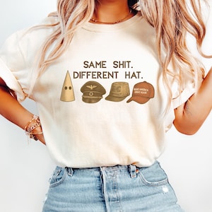 Könnte beinhalten: Cremefarbenes T-Shirt mit dem Text "SAME SHIT. DIFFERENT HAT." und Illustrationen von vier verschiedenen Hüten: einer spitz zulaufenden Kapuze, einer Militärmütze, einem Fedora und einer Baseballkappe.