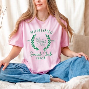 May include: A pink t-shirt with a green Mahjong Social Club logo. The logo features two Mahjong tiles with the text "EST. 2025" and "Olivia".