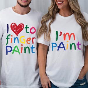 Może przedstawiać: Dwie białe koszulki z kolorowym tekstem. Jedna koszulka mówi "I ❤️ to finger paint" z czerwonym sercem. Druga mówi "I'm paint" w tęczowych kolorach. Obie koszulki są z krótkim rękawem.
