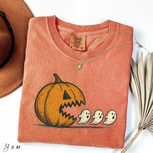Puede incluir: Camiseta naranja Comfort Colors con diseño de Halloween. El gráfico muestra una calabaza tallada comiendo tres pequeños fantasmas. También se ven un collar dorado y un sombrero marrón. La camiseta tiene un aspecto vintage y lavado.