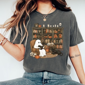 May include: Dark grey t-shirt with a Halloween illustration. A ghost reads a book in an armchair in front of a bookshelf with books, pumpkins, and candles. A black cat sits nearby. Skulls and pumpkins are on the floor.