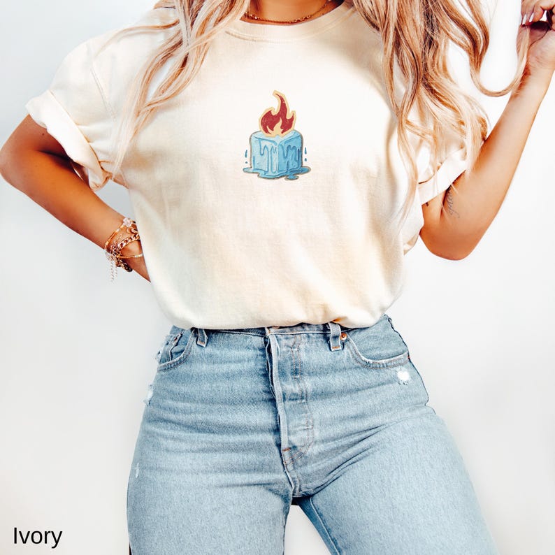 May include: Ivory-colored t-shirt featuring a cartoon illustration of a blue ice cube with a red flame on top. The t-shirt is paired with light blue denim jeans. The word "Ivory" is printed at the bottom left of the image.