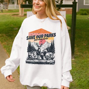 May include: White sweatshirt with a graphic print of a sunset scene with mountains, trees, and animals. The text "Save Our Parks" is printed in a vintage style font with the words "Resist-Defend-Protect" underneath.