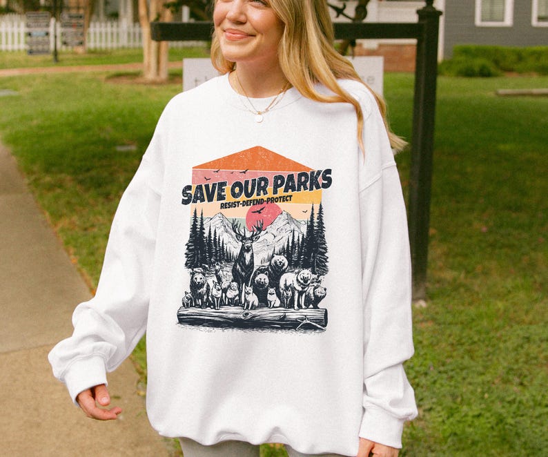 May include: White sweatshirt with a graphic print of a sunset scene with mountains, trees, and animals. The text "Save Our Parks" is printed in a vintage style font with the words "Resist-Defend-Protect" underneath.