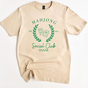 May include: A beige t-shirt featuring a green graphic design that reads "Mahjong Social Club Olivia" with a laurel wreath and two mahjong tiles.