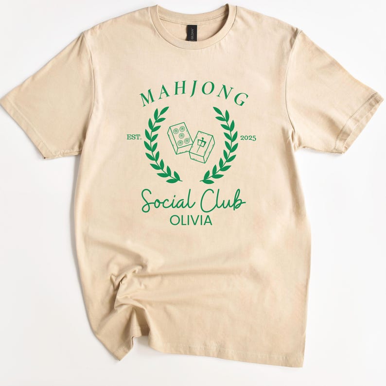 May include: A beige t-shirt featuring a green graphic design that reads "Mahjong Social Club Olivia" with a laurel wreath and two mahjong tiles.
