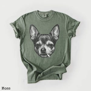 May include: Moss green t-shirt with a black and white illustration of a Chihuahua smoking a cigarette. The shirt has a crew neck and short sleeves. The word "Moss" is printed at the bottom. The t-shirt is made of a soft material.