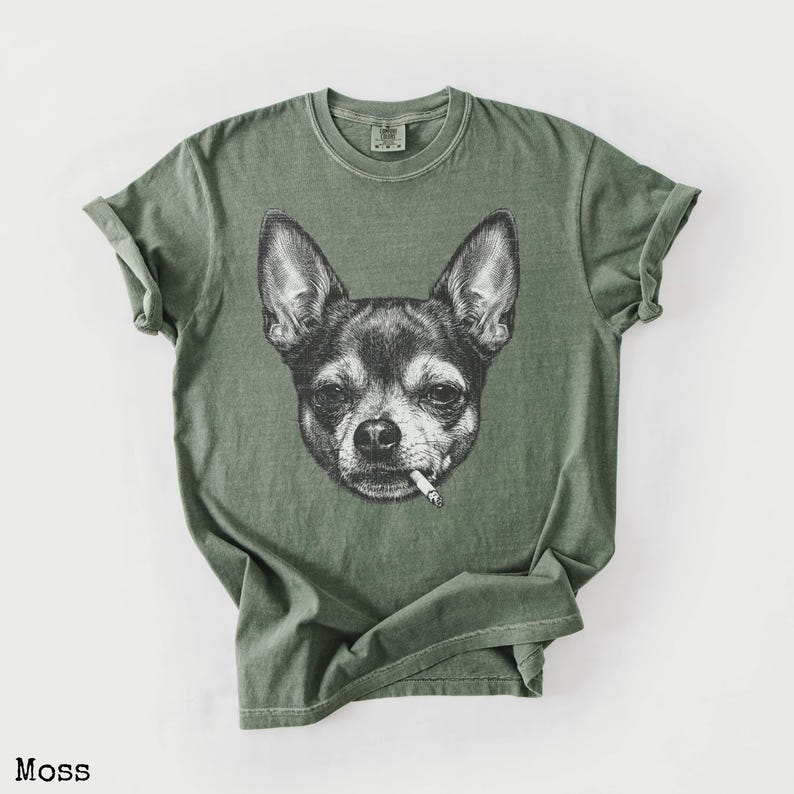 May include: Moss green t-shirt with a black and white illustration of a Chihuahua smoking a cigarette. The shirt has a crew neck and short sleeves. The word "Moss" is printed at the bottom. The t-shirt is made of a soft material.