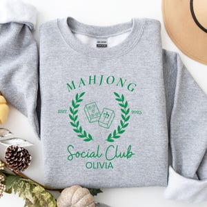 May include: Grey sweatshirt with a green graphic design that says "Mahjong Social Club Olivia" with a wreath and two mahjong tiles. The tiles have a green outline and the text "EST. 2025" is below the wreath.