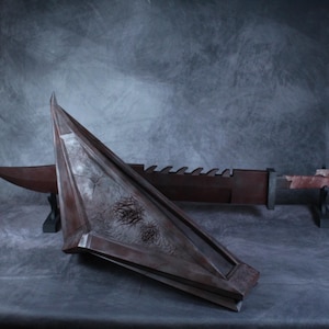 May include: A large, dark brown, triangular pyramid-shaped helmet with a weathered, textured surface and a long, serrated-edge sword featuring a rectangular guard and a wrapped handle, both displayed on black stands against a gray backdrop.