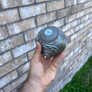 May include: A detailed, metallic sphere with intricate patterns and a central emblem. The sphere is silver and gold in colour, with a textured surface. The design includes circular and linear elements, creating a complex visual texture.