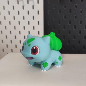 May include: A light blue Bulbasaur figurine with green spots and a green bulb on its back. The figure has red eyes and white feet. The figurine is on a white surface.