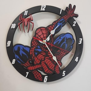 Può includere: Un orologio da parete rotondo con un design di Spider-Man rosso e blu. L'orologio ha una cornice nera con numeri e lancette bianche. Un ragno rosso è attaccato al quadrante. L'orologio è montato su una parete bianca.