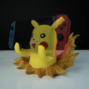 Könnte beinhalten: Ein gelber Pikachu-Themen-Nintendo-Switch-Konsolenständer. Der Ständer zeigt eine lächelnde Pikachu-Figur mit ausgestreckten Armen, einer roten Wange und einer flammenartigen Basis. Ein roter und schwarzer Switch-Controller ist hinter der Figur zu sehen.