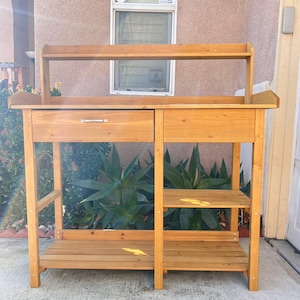 May include: A wooden potting bench with a shelf, a drawer, and two lower shelves. The bench is made of natural wood and has a rustic look.