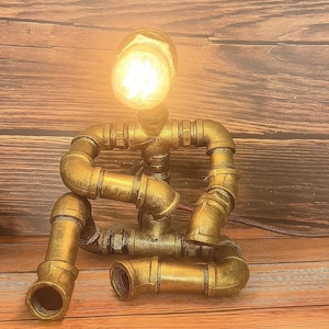 May include: A unique, handcrafted desk lamp in the shape of a person, constructed from brass pipes and fittings. The lamp features a glowing bulb for a head, and is posed in a seated position. The lamp is a decorative accent piece.
