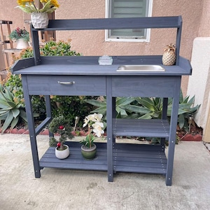 May include: A grey wooden potting bench with a sink, a shelf, and a drawer. The bench has a grey wooden top and a grey metal sink. There are two shelves below the sink and a drawer below the top. The bench is sitting on a concrete patio.
