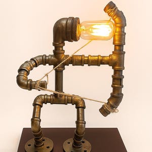 May include: A unique industrial-style lamp made from bronze-colored pipes and fittings. The lamp features a warm, glowing Edison bulb and is mounted on a dark brown wooden base. The design resembles a stylized human figure.