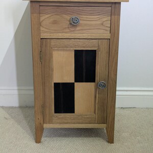 May include: A wooden nightstand with a drawer and a cabinet door. The cabinet door features a checkerboard design with alternating black and light wood squares. The top of the nightstand has a decorative inlay. The drawer and door have spiral-shaped metal knobs.