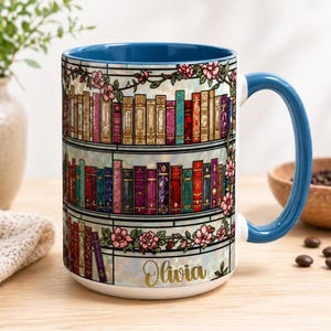 May include: A white ceramic mug with a blue interior and handle, featuring a colorful bookshelf design with pink floral accents. The name "Olivia" is written in gold script on the front.