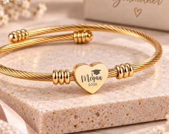 May include: A gold-toned cable wire bracelet featuring a central heart-shaped charm engraved with a graduation cap icon, the name Megan, and the year 2026. The bracelet has decorative ribbed metal accents on either side of the heart charm.