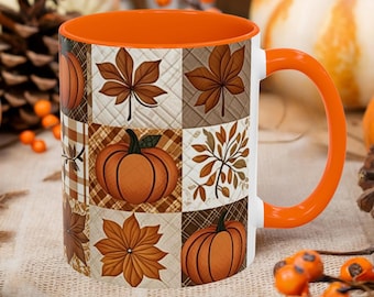 May include: A ceramic mug with a bright orange handle and interior, featuring a patchwork design on the exterior. The pattern consists of alternating squares displaying illustrations of orange pumpkins, brown autumn leaves, and plaid textures.
