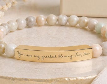 May include: A beaded bracelet featuring smooth, multi-colored pastel stones and a central gold-toned metal bar. The bar is engraved with the cursive text, "You are my greatest blessing. Love, Mom." The bracelet is presented with a branded gift box.