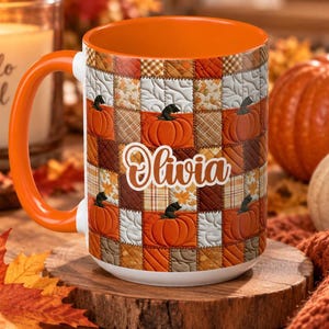 May include: A ceramic mug with an orange handle and interior, featuring a patchwork quilt pattern of orange pumpkins, brown plaid, and cream-colored leaf designs. The name Olivia is printed in white cursive script across the center of the mug.