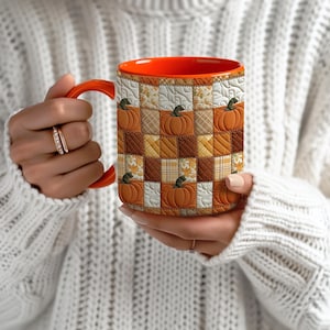 May include: A ceramic mug with a vibrant orange interior and handle, featuring a quilted patchwork pattern on the exterior. The design consists of alternating squares in shades of orange, brown, and cream, decorated with pumpkin and leaf motifs.