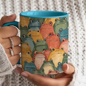 May include: A ceramic mug with a light blue interior and handle, featuring a colorful pattern of cartoon frogs in various shades of green, yellow, orange, and blue. The mug is held by a person wearing a white sweater.