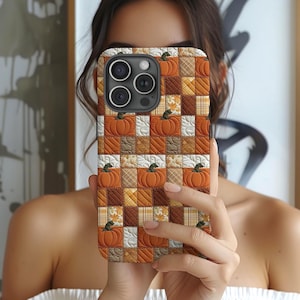 May include: A smartphone case featuring a quilted patchwork design with alternating squares of orange pumpkins, brown textures, and cream-colored plaid patterns. The case has a cutout for a triple-lens camera system on the upper left side.