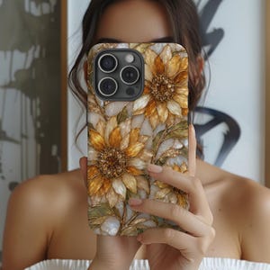 以下が含まれることがあります： 金、黄、白の花びらを持つヒマワリの模様が描かれた長方形のスマートフォンケース。花の中心部は暗い色でキラキラとしたアクセントがあり、緑の葉が添えられています。背景は真珠のような光沢のある大理石風のデザインです。
