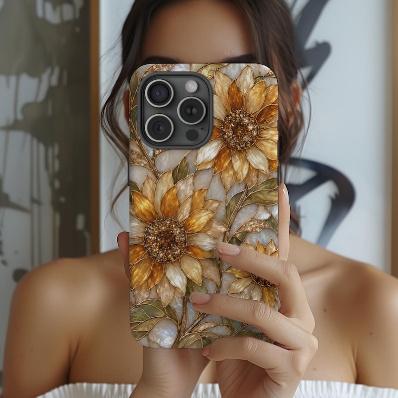 以下が含まれることがあります： 金、黄、白の花びらを持つヒマワリの模様が描かれた長方形のスマートフォンケース。花の中心部は暗い色でキラキラとしたアクセントがあり、緑の葉が添えられています。背景は真珠のような光沢のある大理石風のデザインです。