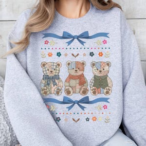 May include: A light gray sweatshirt featuring three patchwork teddy bears with blue bows and floral accents. The bears have different patterned fabrics and wear scarves and vests. The design is centered on the chest.