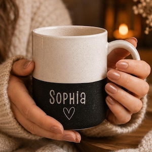 May include: A two-toned ceramic mug featuring a speckled cream-colored upper section and a solid black lower section. The name Sophia is engraved in white lettering on the black portion, accompanied by a small, simple white heart outline below it.