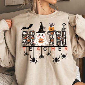 May include: A beige crewneck sweatshirt featuring a graphic print with the text MATH TEACHER. The letters M, T, and H are filled with a pattern of ghosts, pumpkins, and candy corn. A ghost in a witch hat, a black cat, and a skeleton are included.