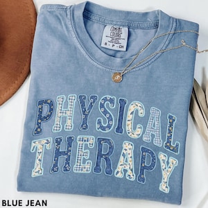 May include: A blue jean Comfort Colors t-shirt with the words "PHYSICAL THERAPY" in a floral and plaid design. The shirt has a crew neck and short sleeves. A gold necklace with a pendant is draped across the shirt.
