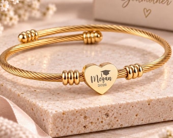 May include: A gold-toned cable wire bangle bracelet featuring a central heart-shaped charm. The charm is engraved with a graduation cap icon, the name Megan, and the year 2026. The bracelet has decorative metal beads on each side of the heart.