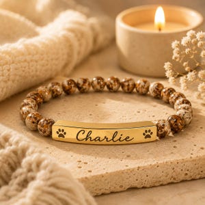 May include: A beaded bracelet with brown and cream-colored stones. The gold-colored bar in the center is engraved with the name "Charlie" and paw prints. A lit candle and decorative flowers are in the background.
