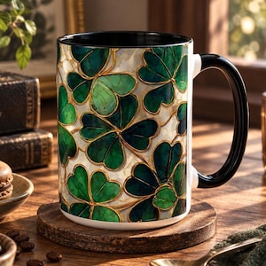 May include: A ceramic mug featuring a repeating pattern of green shamrocks with gold outlines against a white marble-like background. The mug has a solid black interior and a matching black handle, providing a high-contrast look for this drinkware.